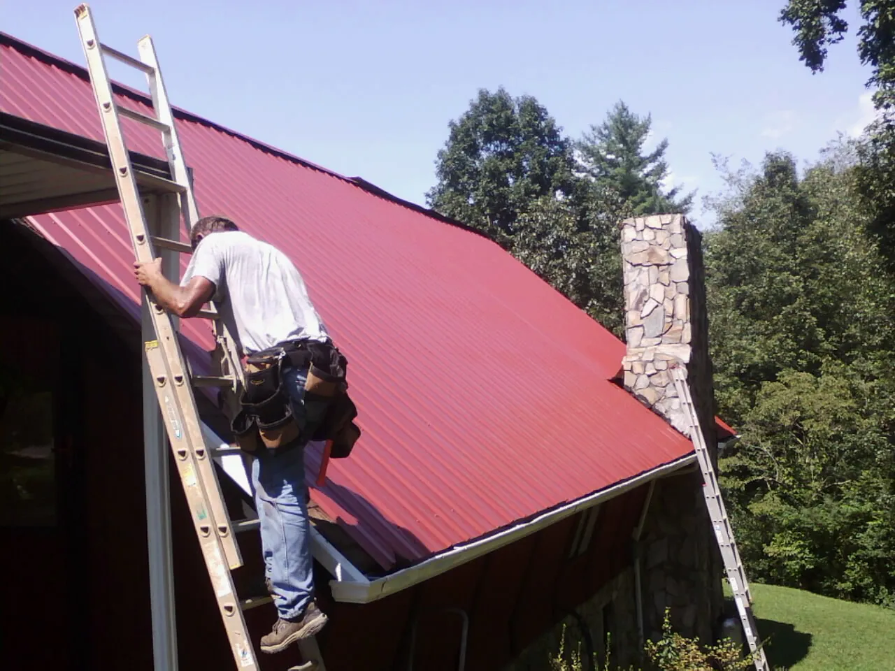 Roofer on a ladder beside a red metal roof and stone chimney — Soffit & Fascia Repair by Roofing Brothers in Buffalo Grove